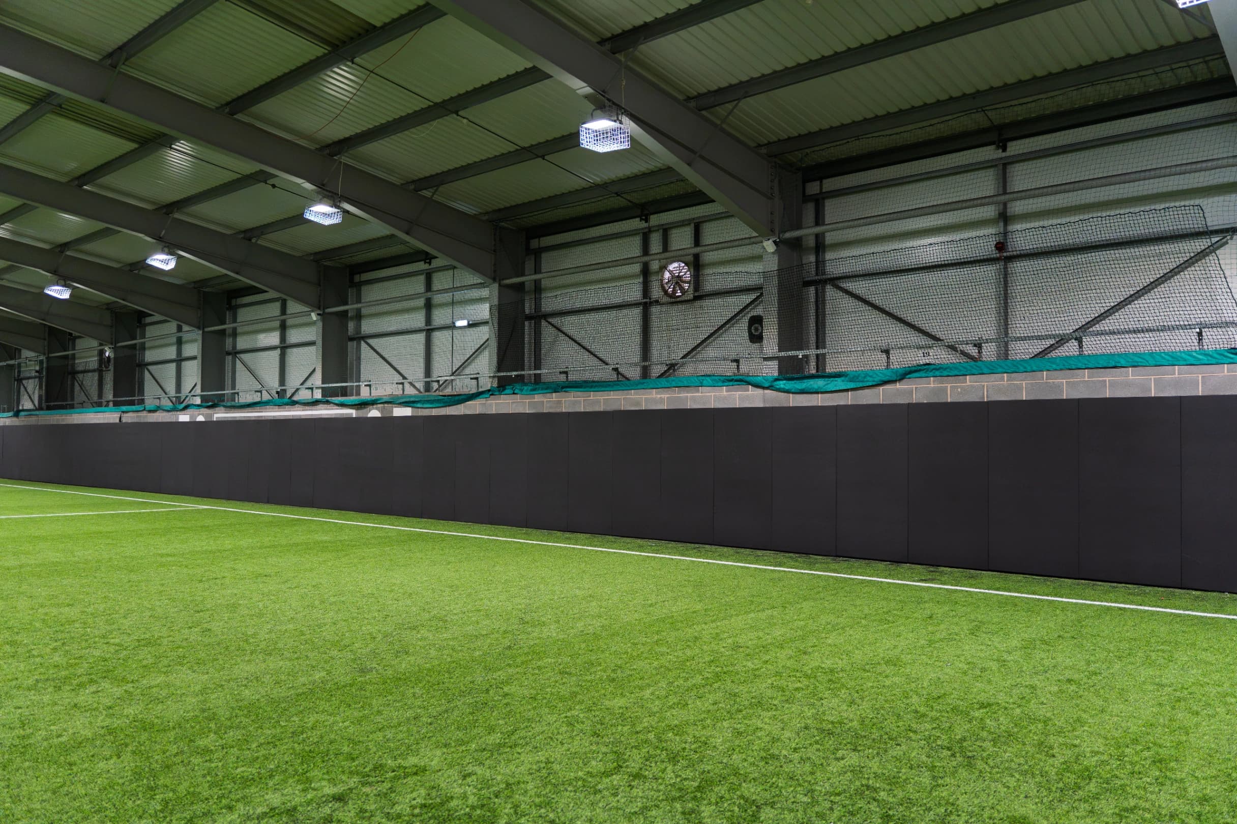 Black sports pitch safety padding made from heavy duty PVC covers mounted along an indoor turf pitch wall beneath metal roof beams and lights.