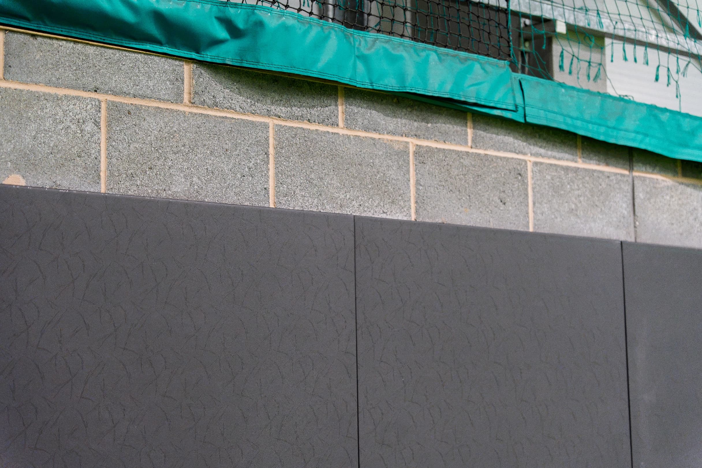 Dark grey sports pitch safety padding panels made from heavy-duty PVC mounted on a concrete block wall beneath green netting.