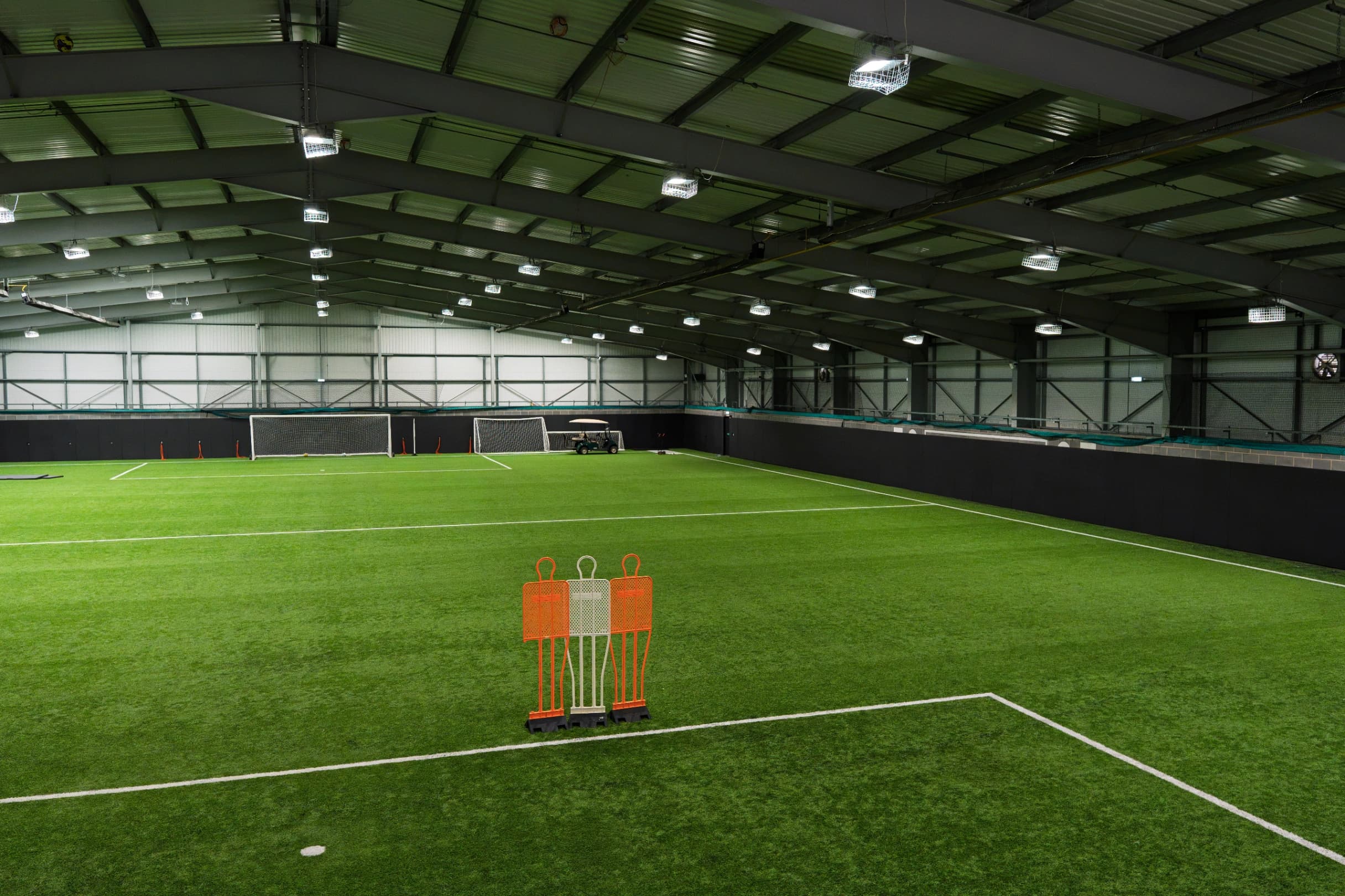 Indoor sports pitch with green artificial turf, perimeter safety padding along the walls, three training mannequins in the foreground and goals at the far end.
