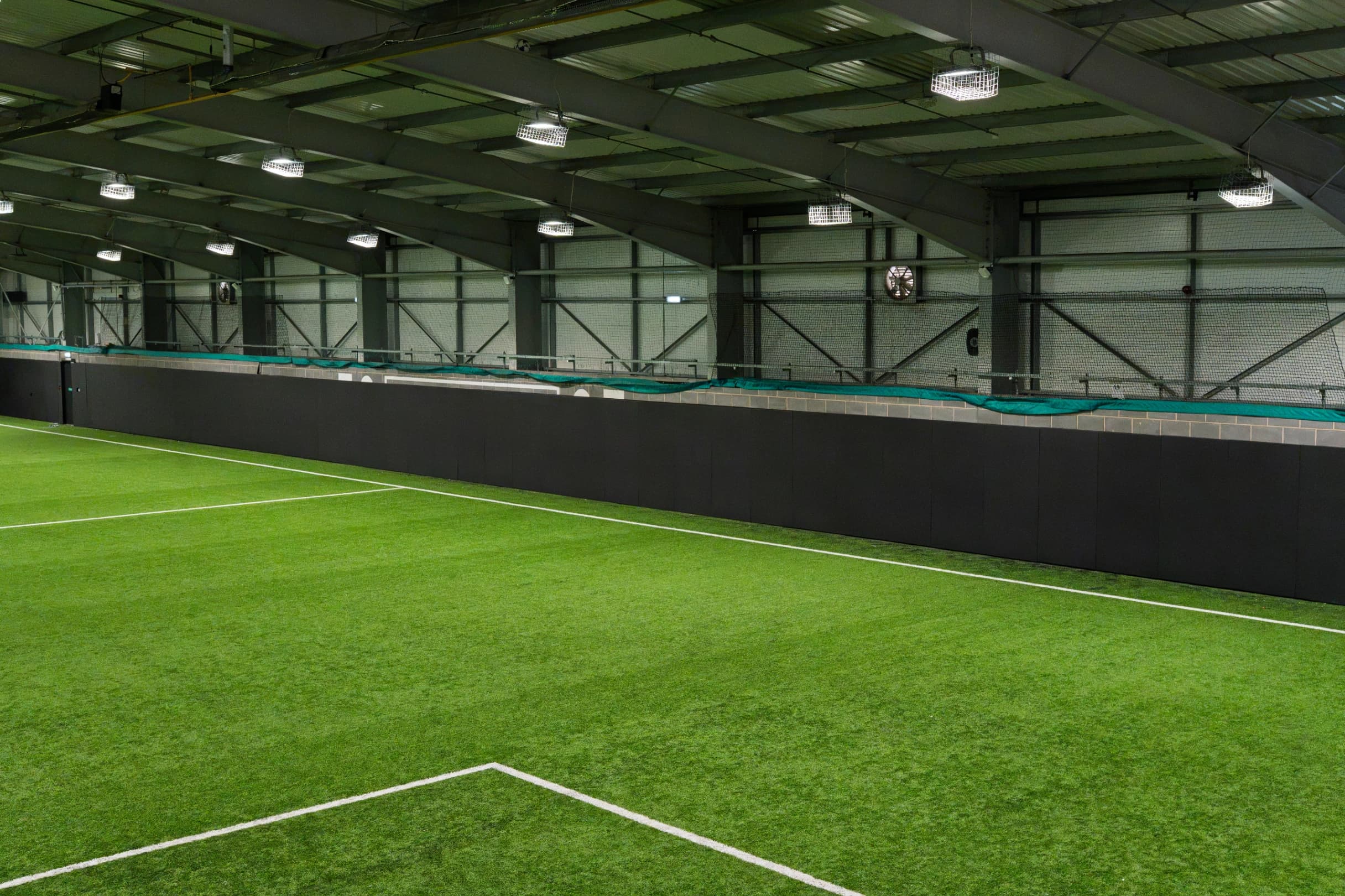 PVC-covered sports pitch safety padding panels lining an indoor artificial turf pitch wall beneath steel roof trusses and overhead lights.