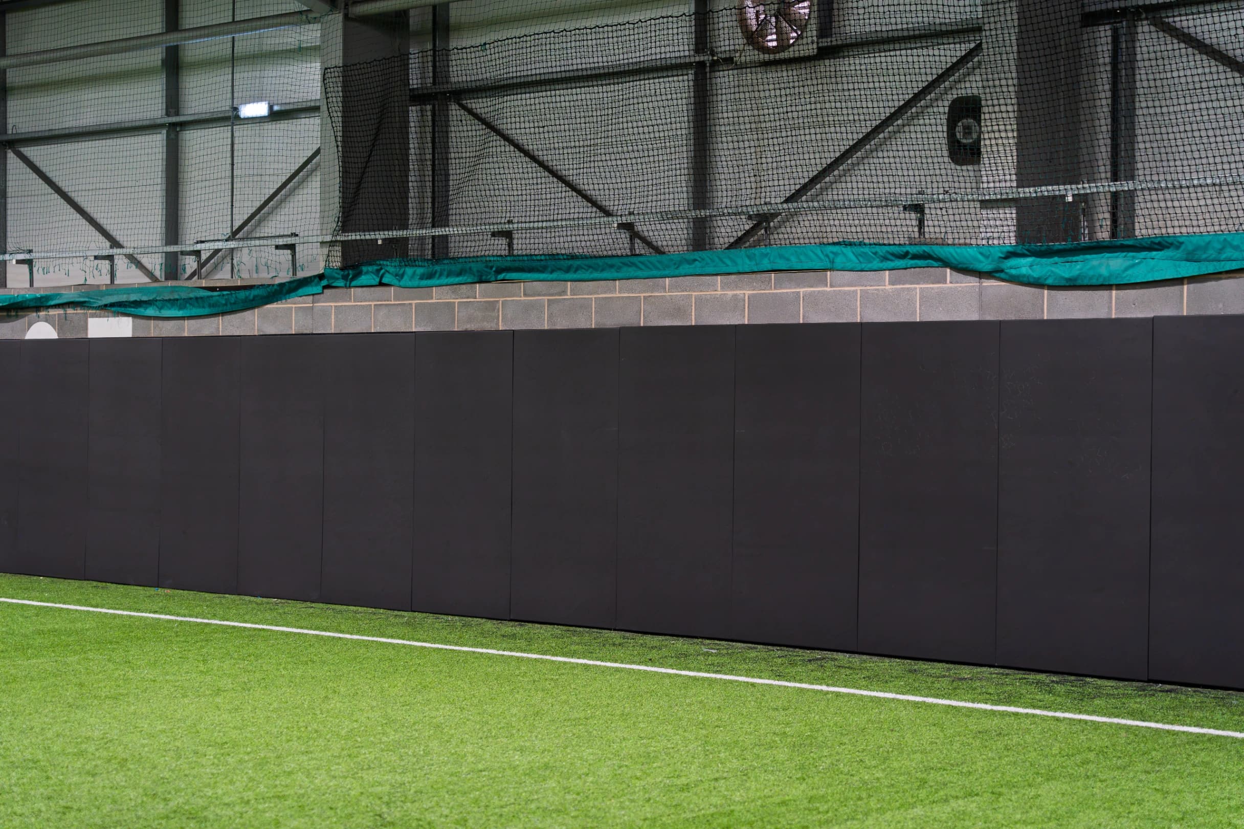 Black sports pitch safety padding panels made from heavy duty PVC lining an indoor wall above green artificial turf.