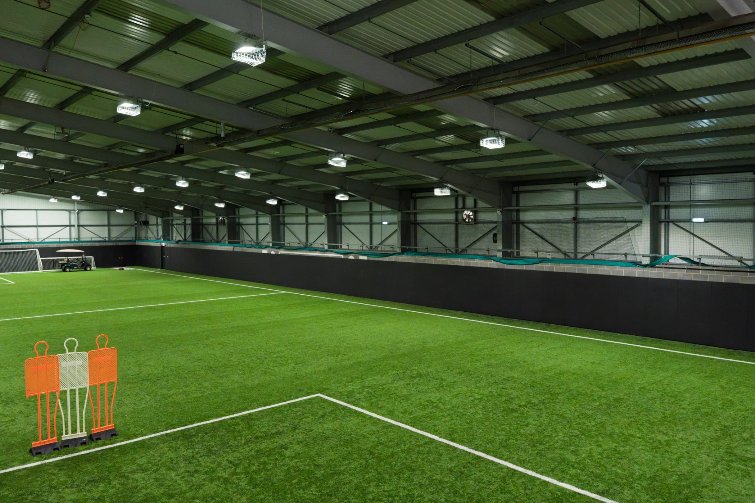 Black sports pitch safety padding lining the perimeter wall of an indoor artificial-turf football pitch beneath steel roof beams and lights.