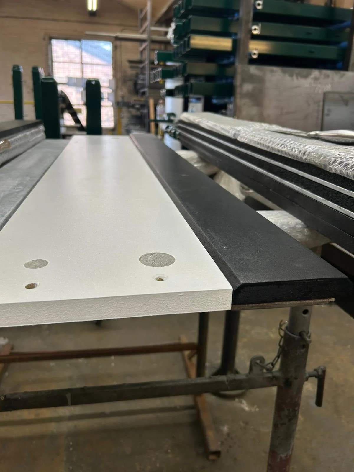 White wooden take-off board with pre-drilled holes and a black rubber edge resting on galvanized steel supports in a workshop.